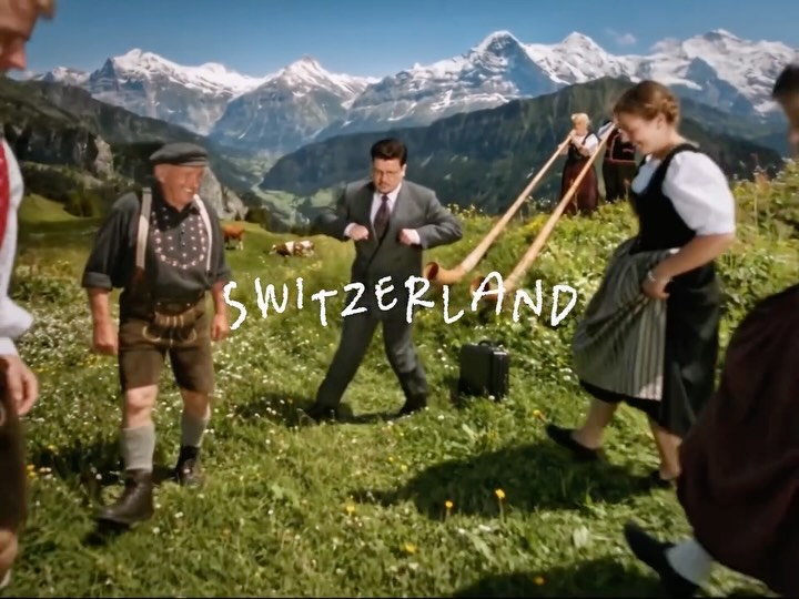 Still from the series He doesn't stop: the man in a grey suit dancing on an alpine meadow with people in Swiss folk costume and alphorns.